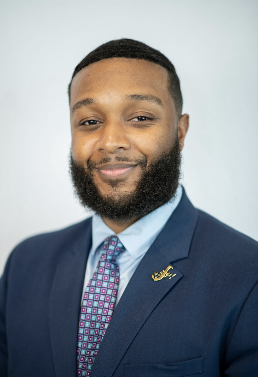 Kiante Bush with beard in navy suit with purple tie and baby blue shirt before grey background.