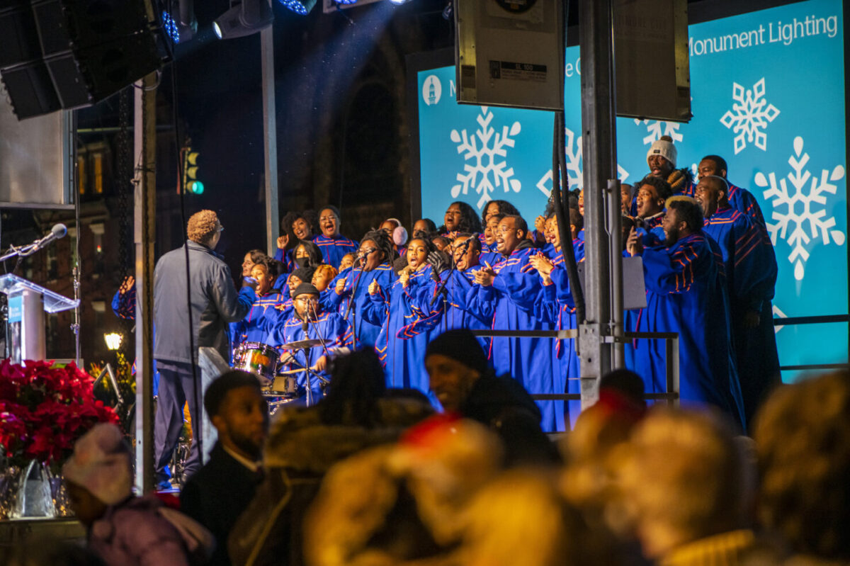 Choir members in blue and orange robes perform in front of a light blue screen with white snowflakes.