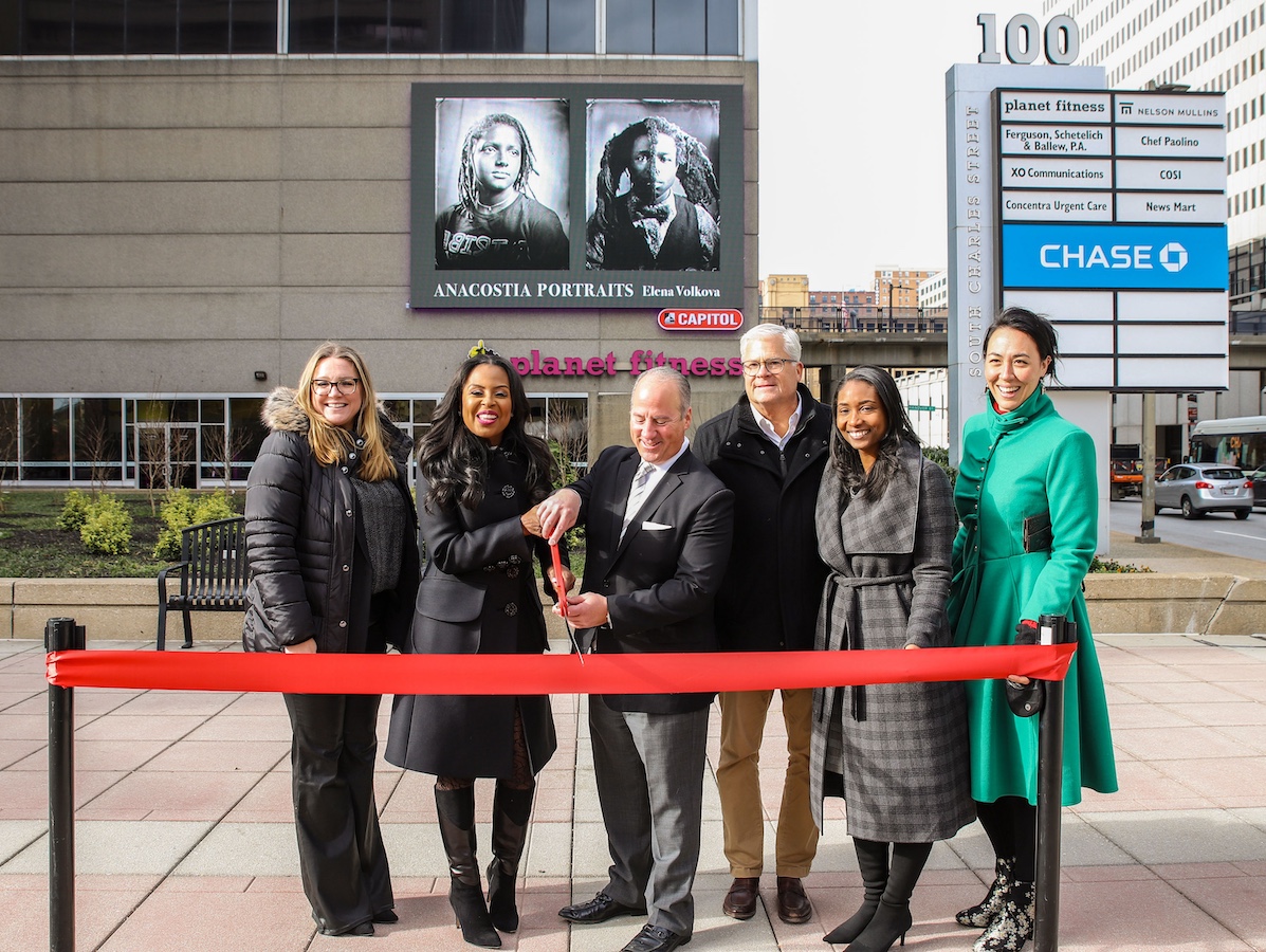 A group of people standing behind a red ribbon in front of digital billboard.