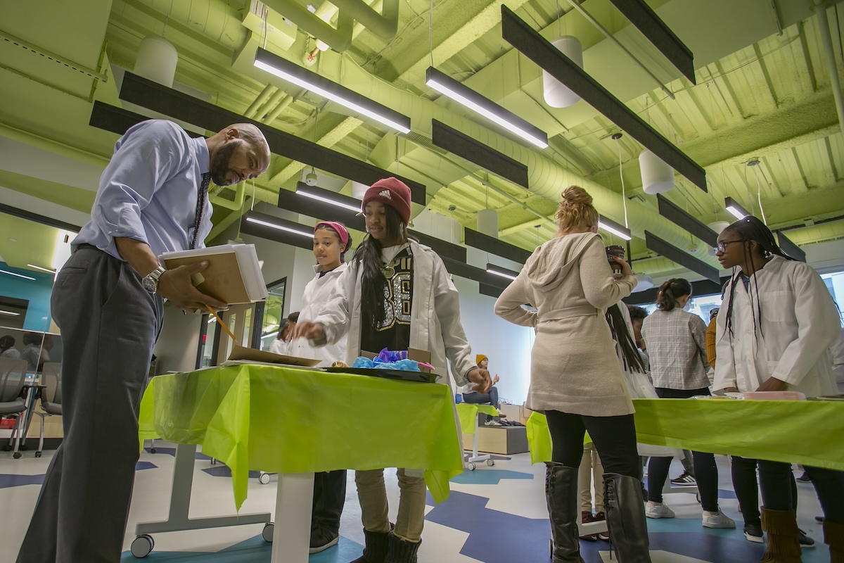 A group of young people in lab coats standing around tables in a room.