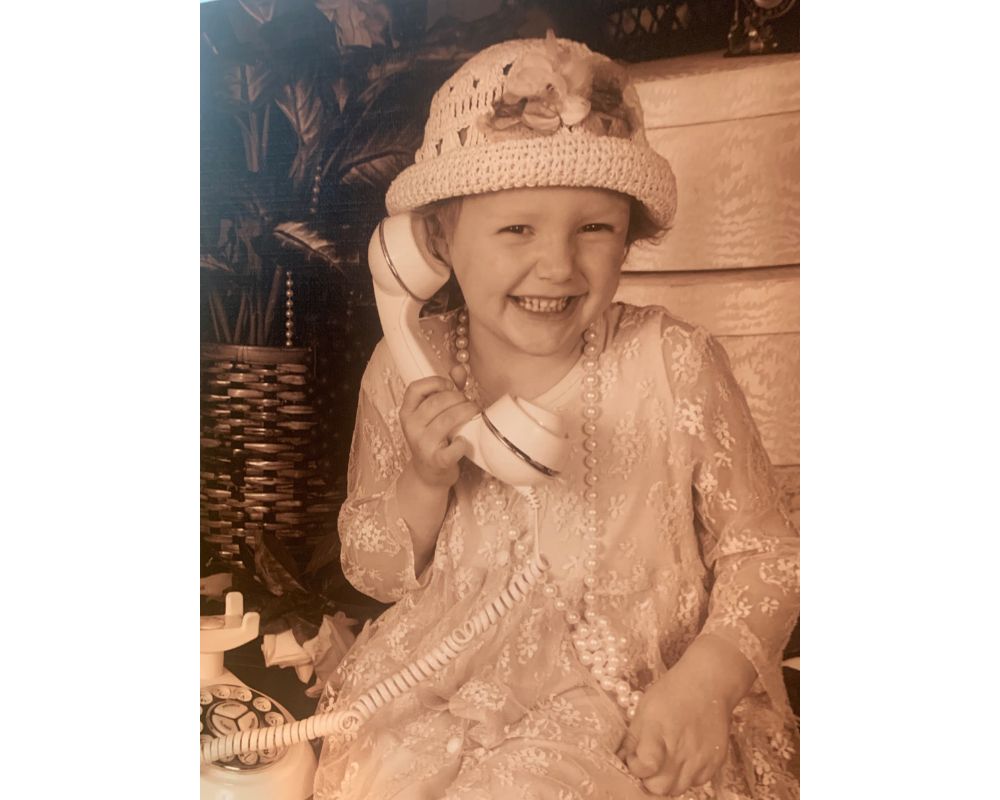 Sepia-toned photo of Pava LaPere as a young girl in white dress with pearls and hat. 