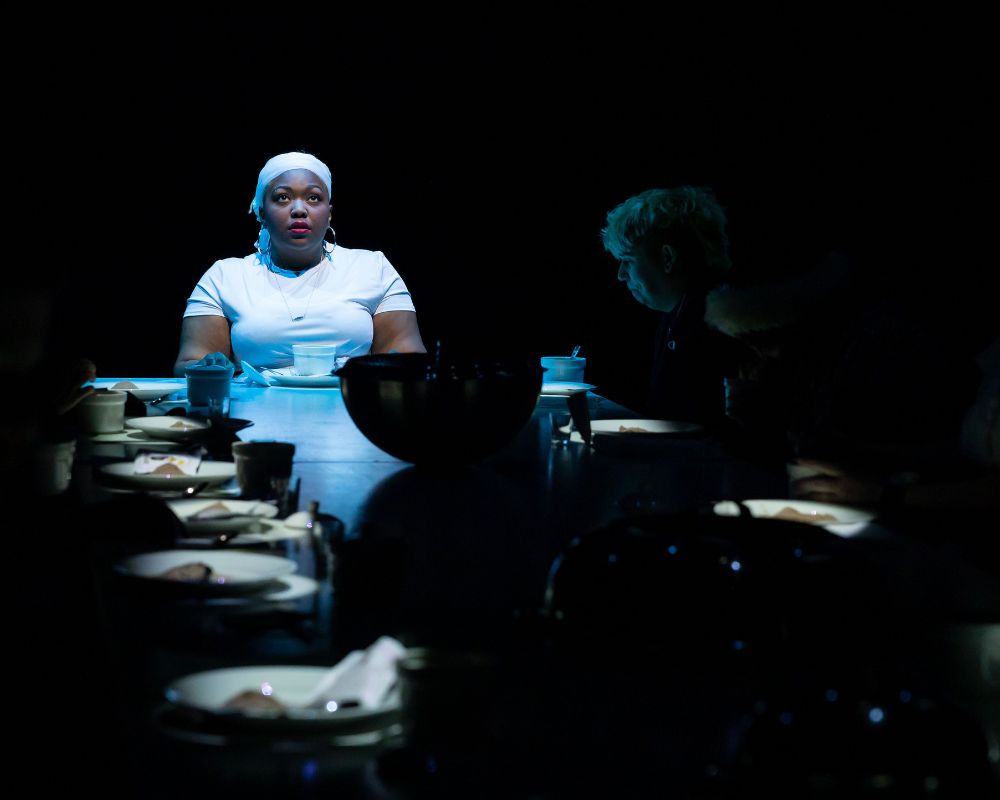 A woman in white sits at a table surrounded by people in silhouette 