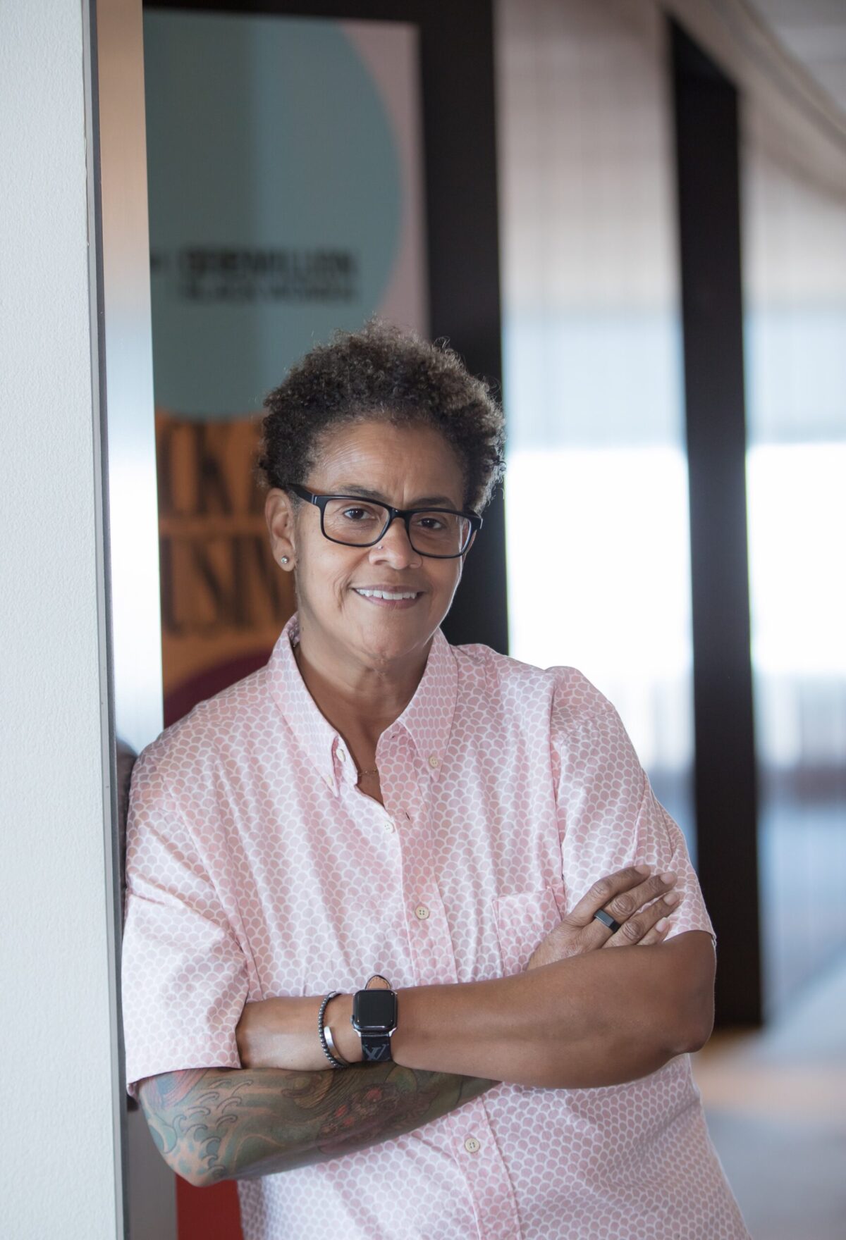 A woman wearing glasses and a pink shirt leaning against a wall.