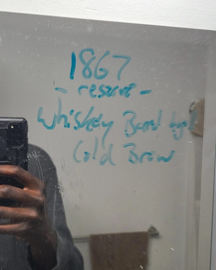 A man's picture of a mirror with blue text reading "1867 Reserve: Whiskey Barrel Aged Cold Brew"