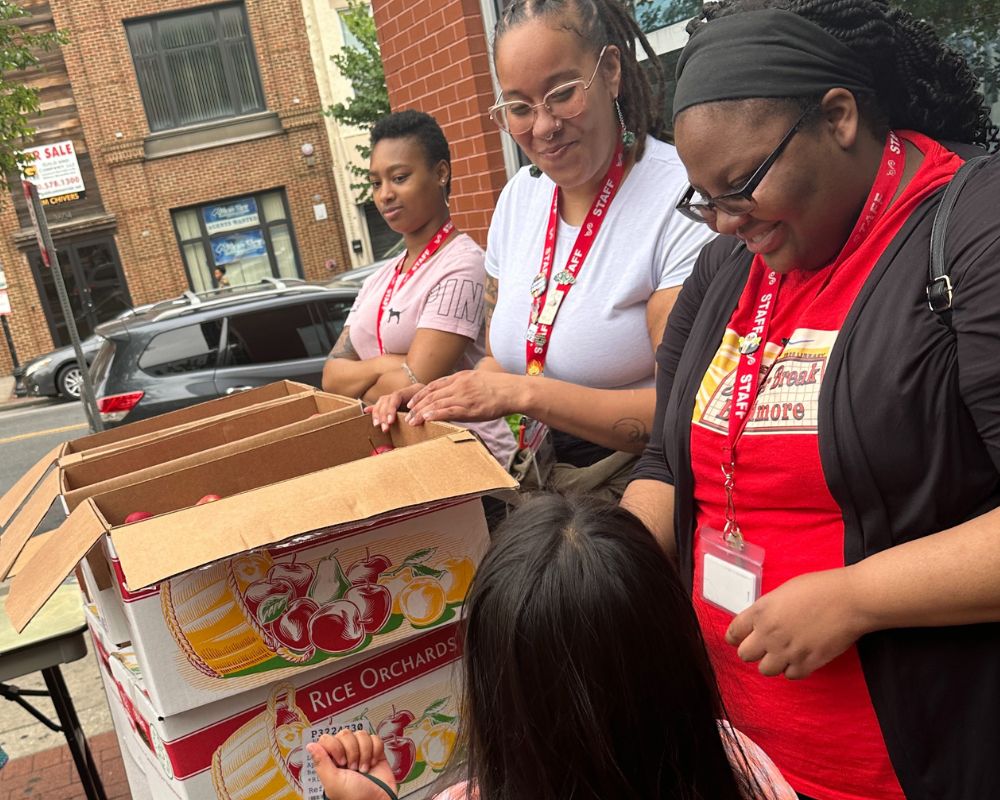 A group of women wearing red lanyards standing next to boxes of food.