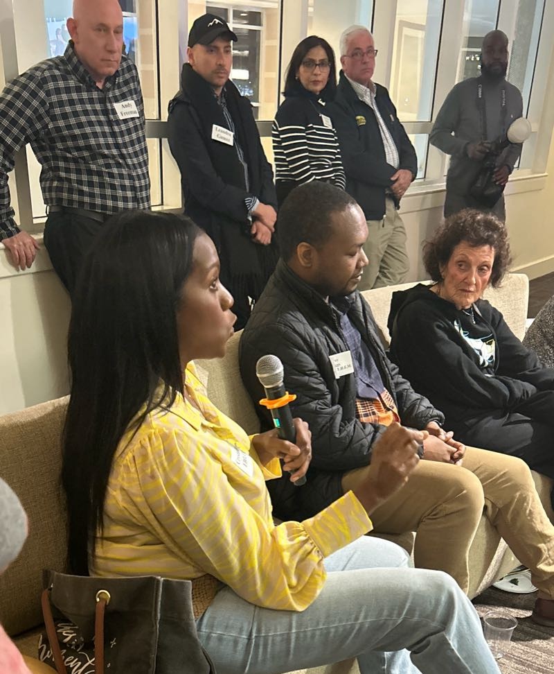 A group of people sitting in a room while a woman in a yellow shirt speaks into a microphone.