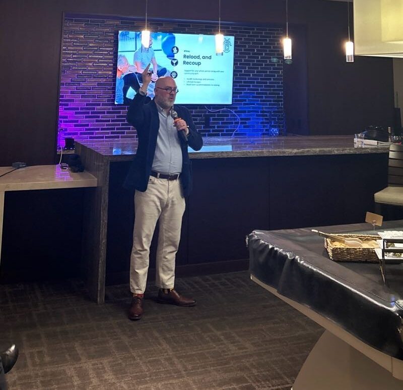 Joel Benge in navy blazer and brown pants standing in front of screen