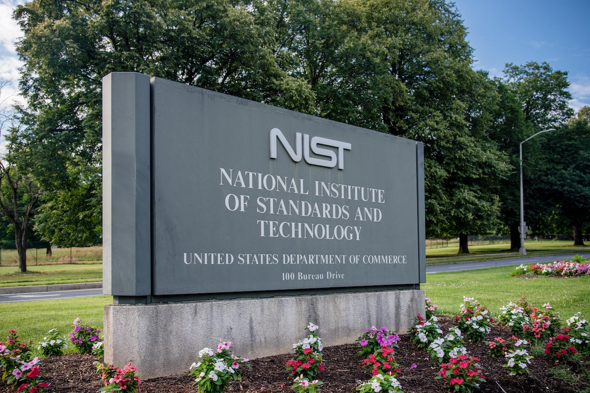 Grey NIST sign with white text near pink flowers by green field.