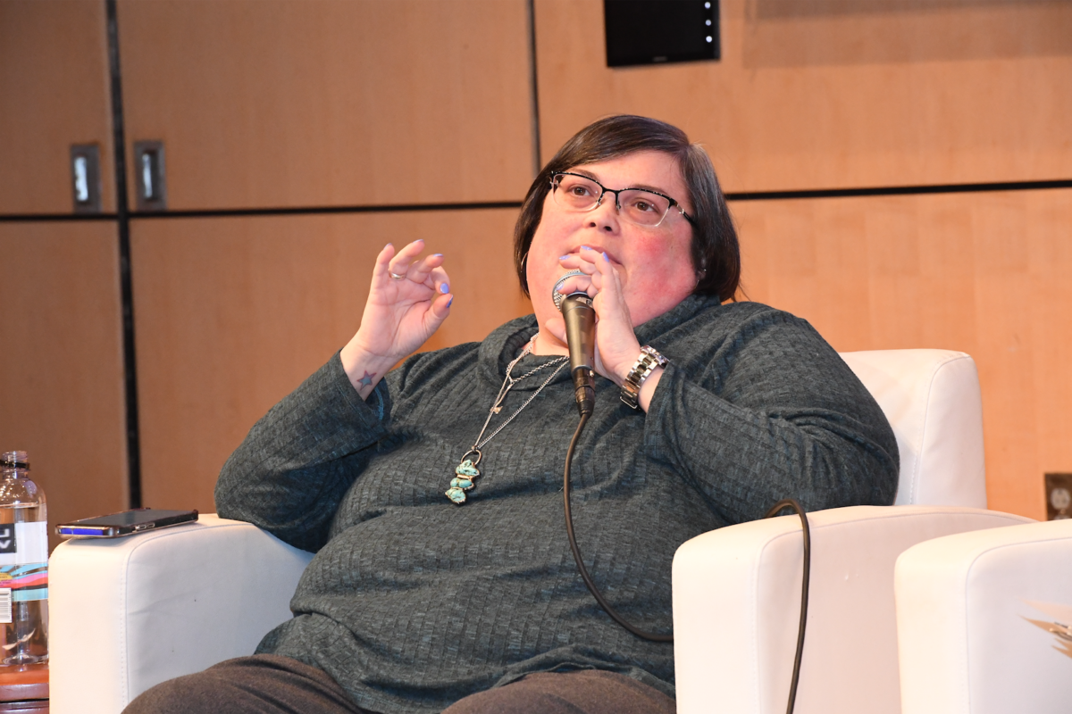 Joan Donovan in grey sweater and brown pants on white chair.