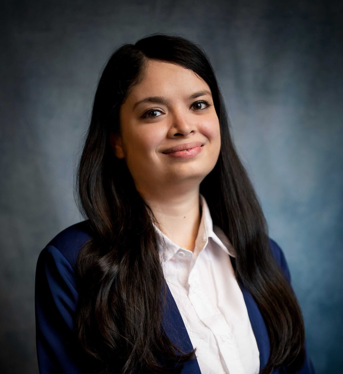 Vishakha Jha in navy blazer and white dress shirt in front of grey-blue background.