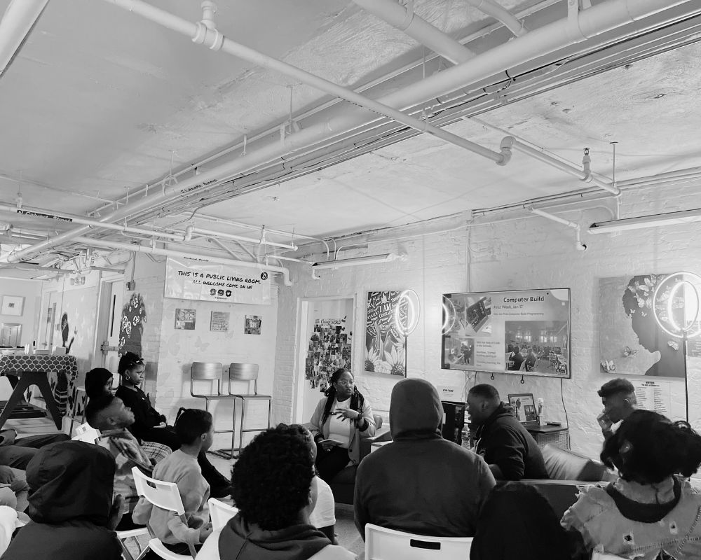 A black and white photo of a group of people sitting in chairs and surrounded by artwork on the walls.