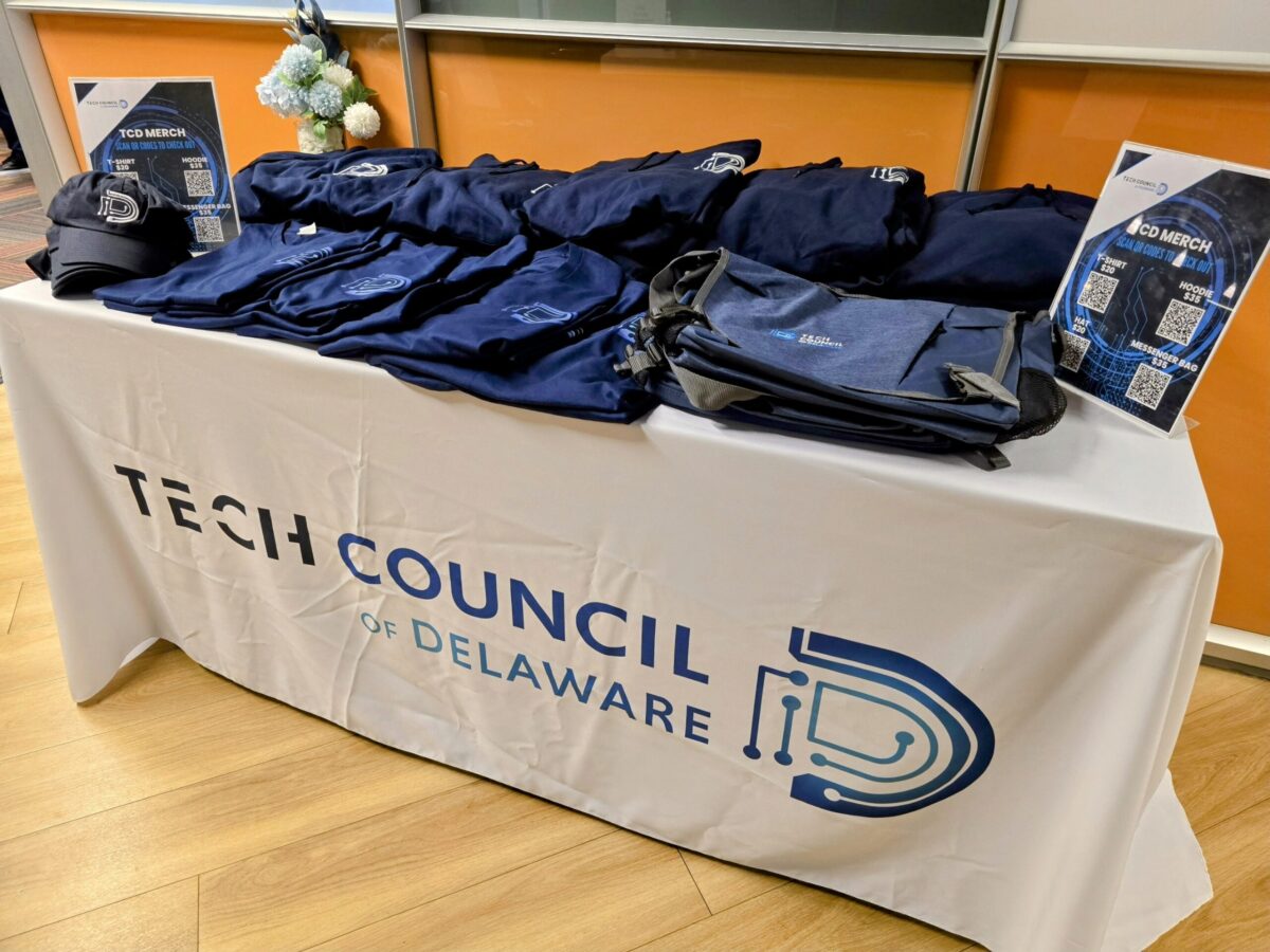 A table draped with a 'tech council of delaware' banner, displaying promotional materials and a stack of navy blue polo shirts.