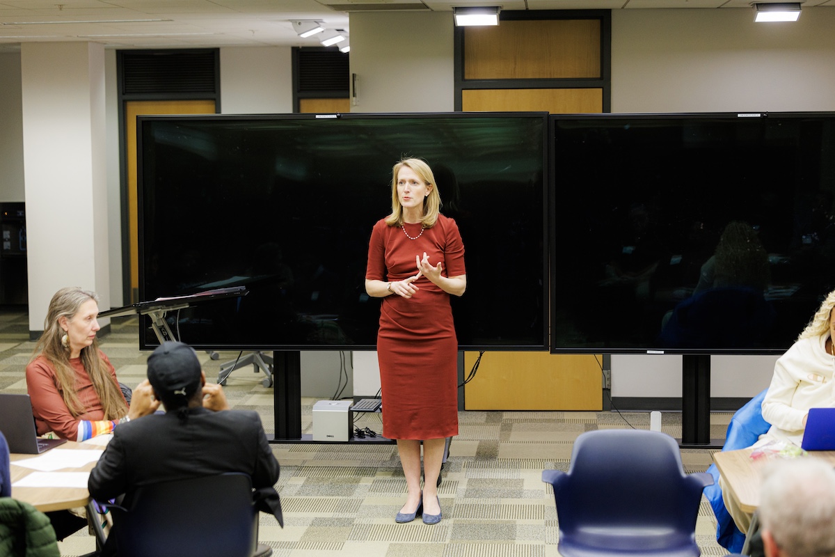 Brooke Lierman in red dress speaking to people at tables in front of two black large TV screens.