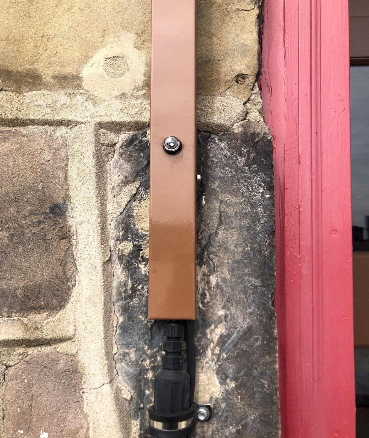 Brown metal door strip secured with a screw between a stone wall and a red door frame.