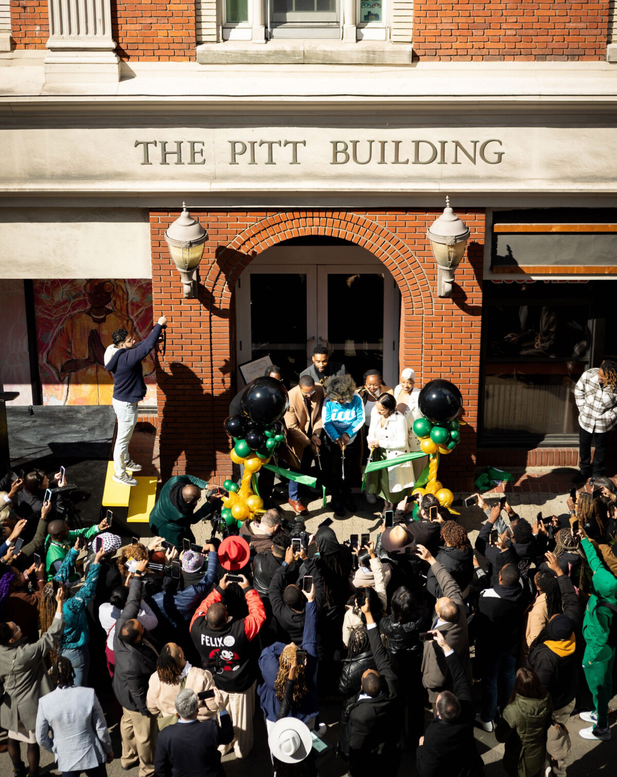 A person addresses a crowd from the entrance of the Emerald City Coworking Space in Pittsburgh, serving Black entrepreneurs, during a daytime event.