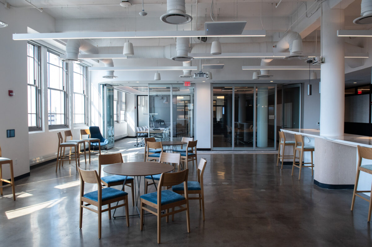 Modern office break room with tables, chairs, and bar-style seating by the windows.