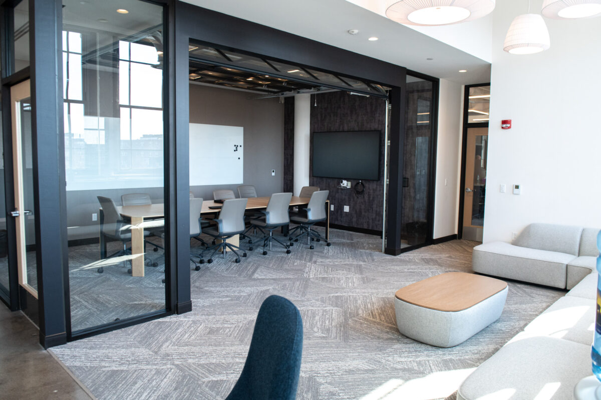 Modern office lounge and meeting room with glass doors, furniture, and a mounted tv.