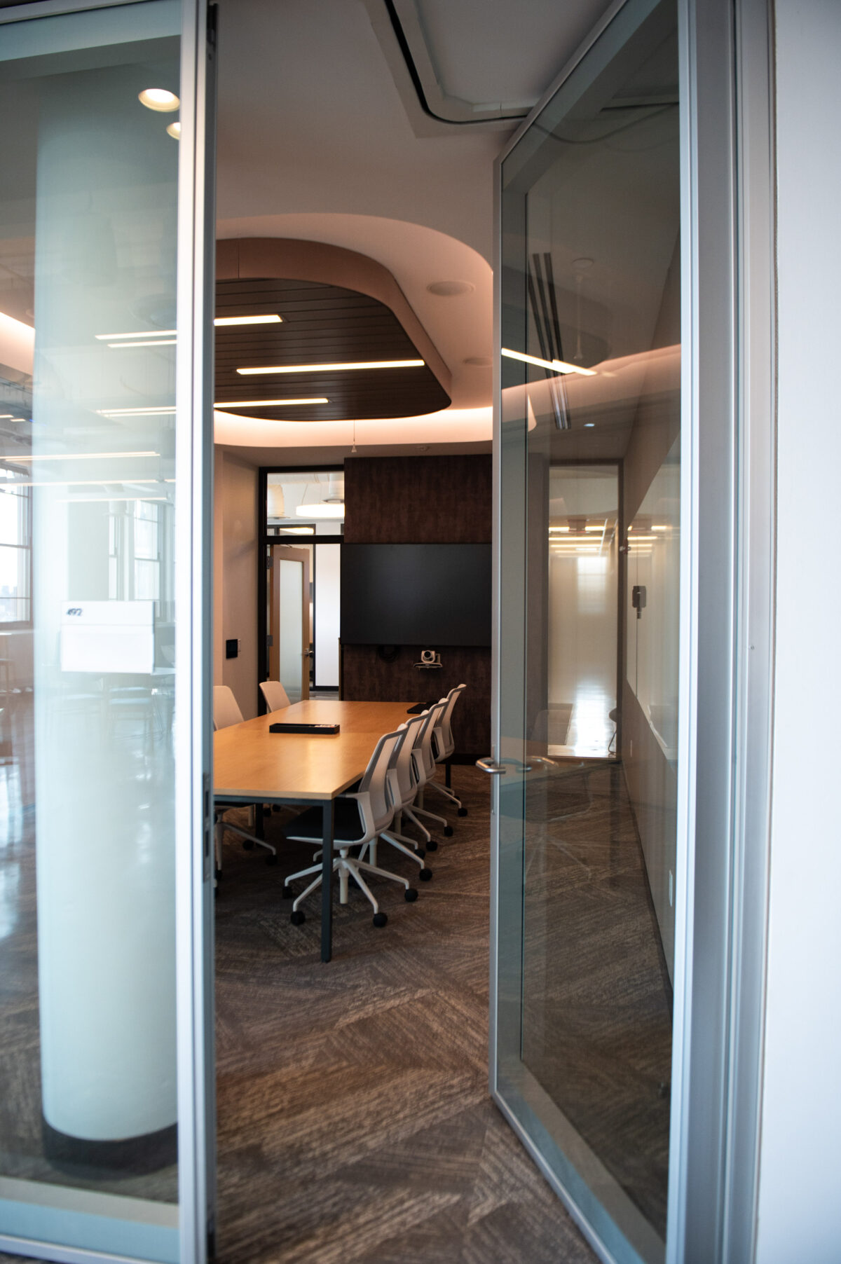 Modern conference room seen through glass doors with a long table and chairs, equipped with a presentation screen.