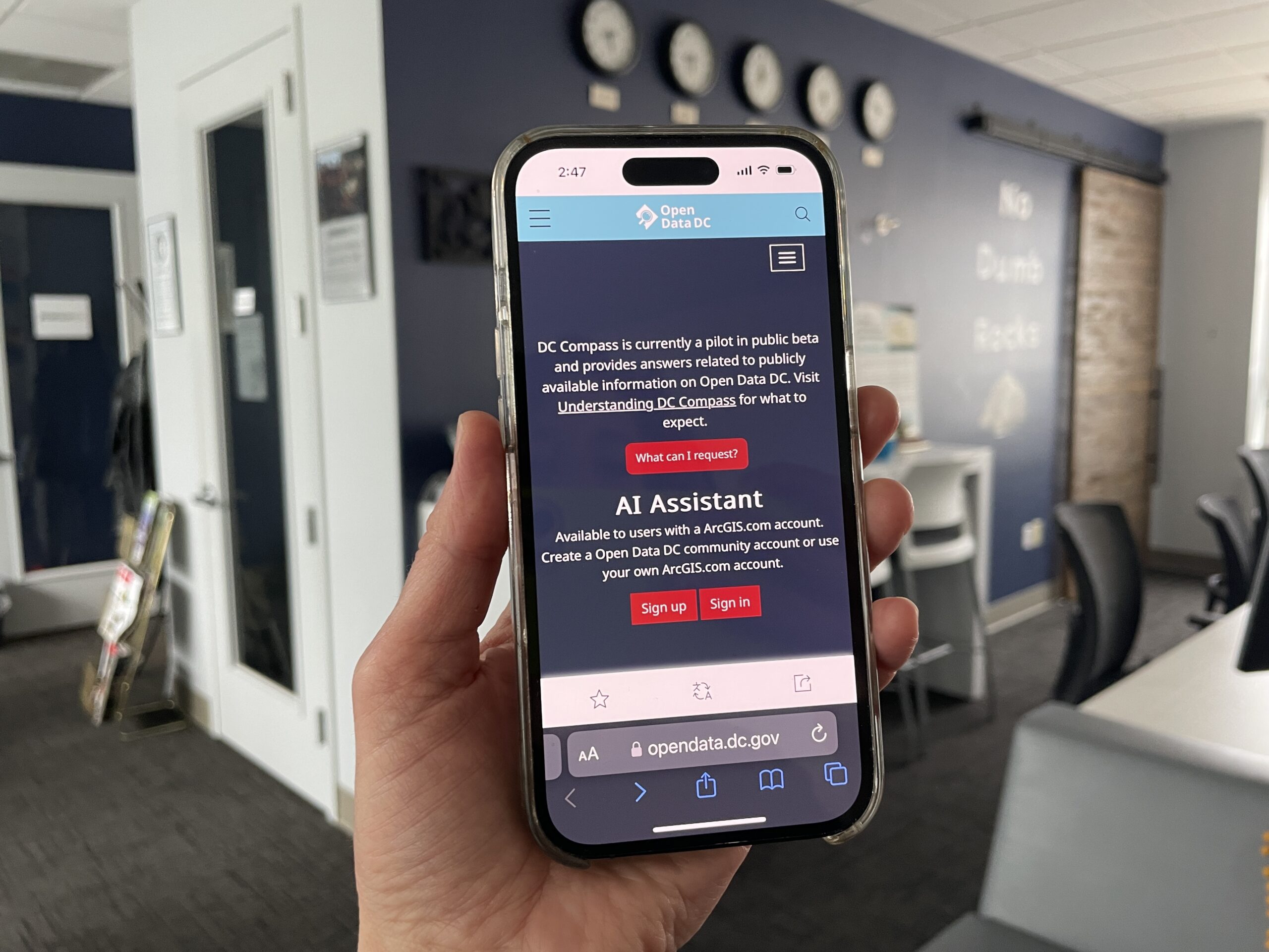 Person holding a smartphone displaying the Open Data DC website with an AI Assistant message. Office setting with a table, chairs, and clocks on the wall in the background.