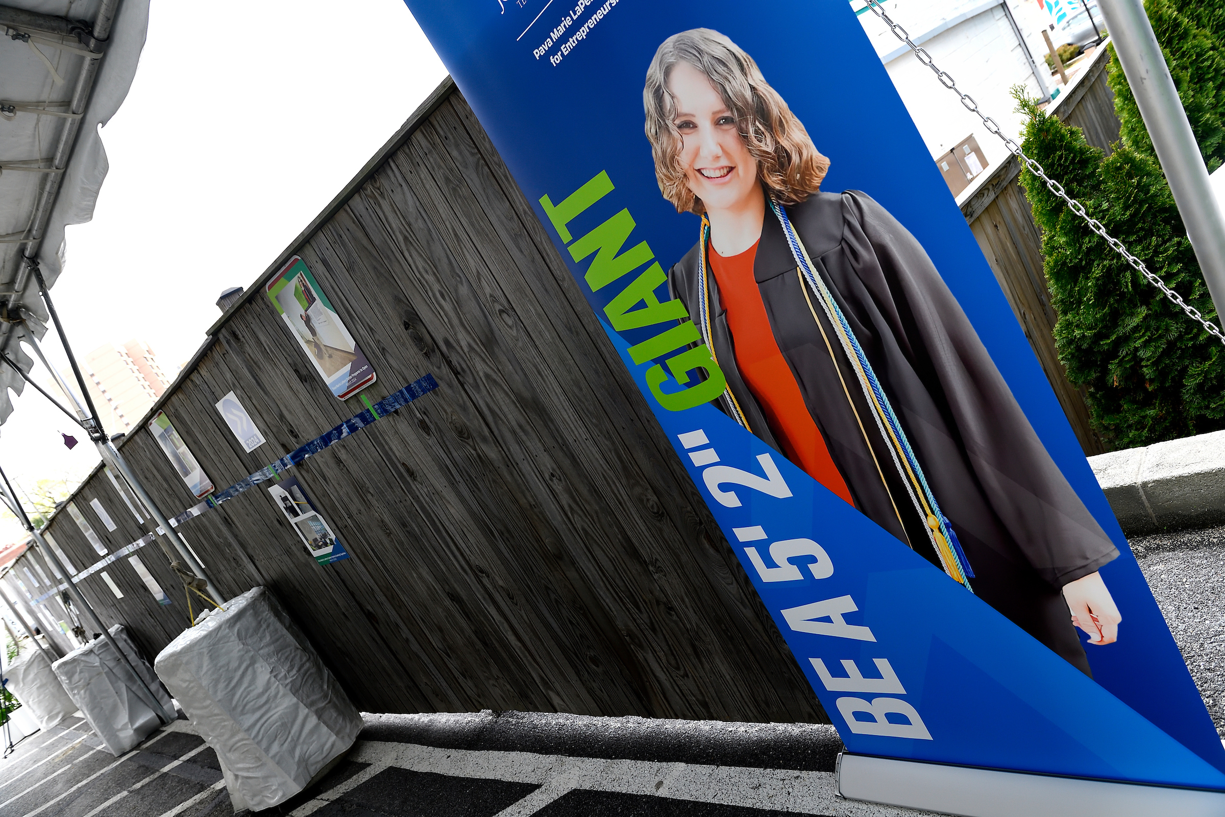 Portrait of Pava LaPere in graduation attire on blue banner near brown wood fence.