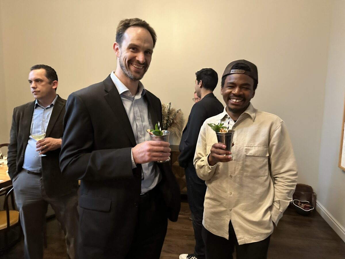Two men smiling and holding drinks at a social event, with other guests chatting in the background.