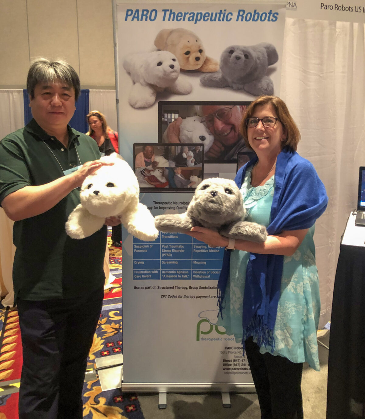 Two individuals presenting paro therapeutic robot seals at a conference booth.