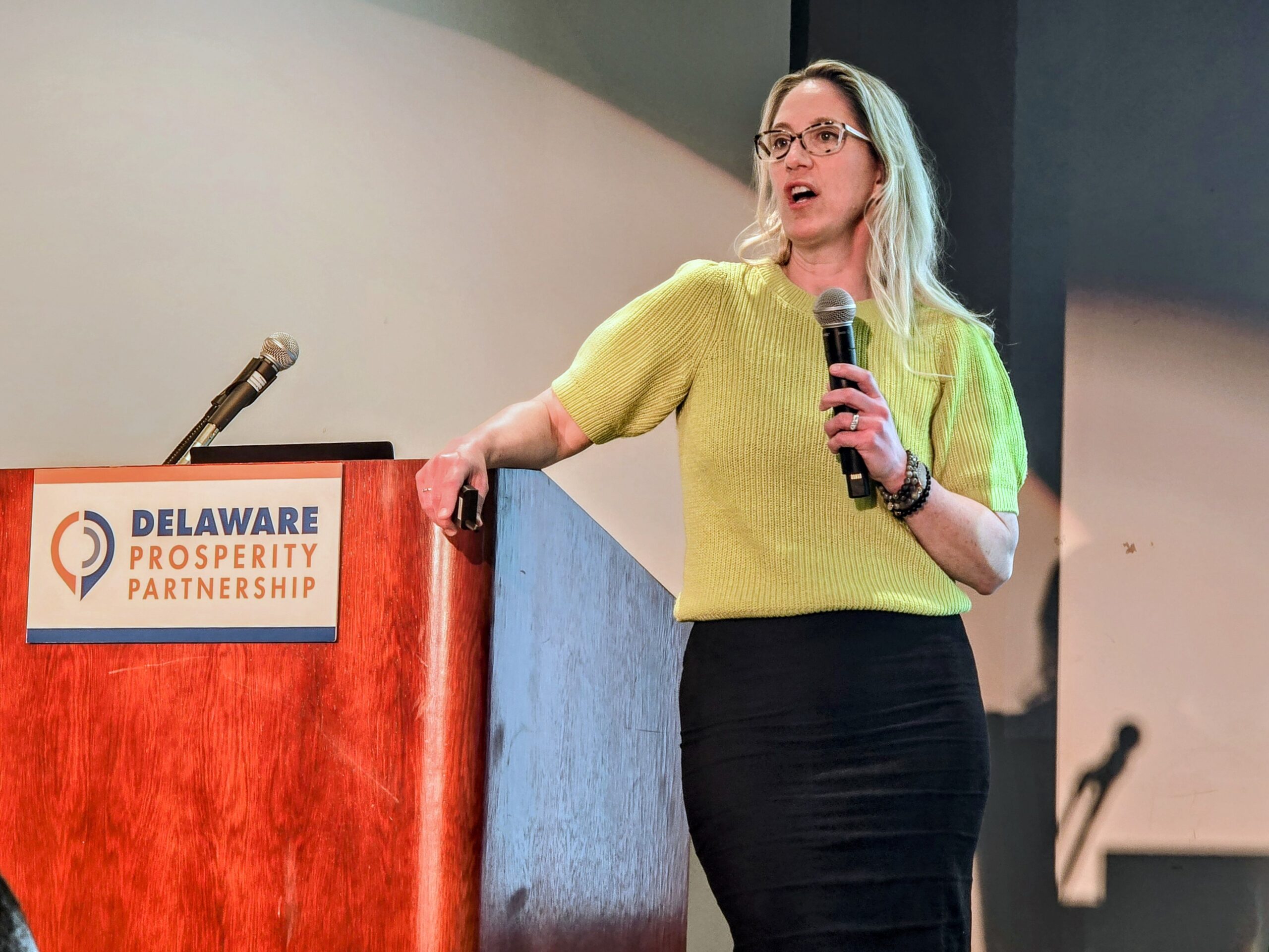 A person stands at a podium labeled "Delaware Prosperity Partnership," speaking into a microphone while holding a remote clicker.