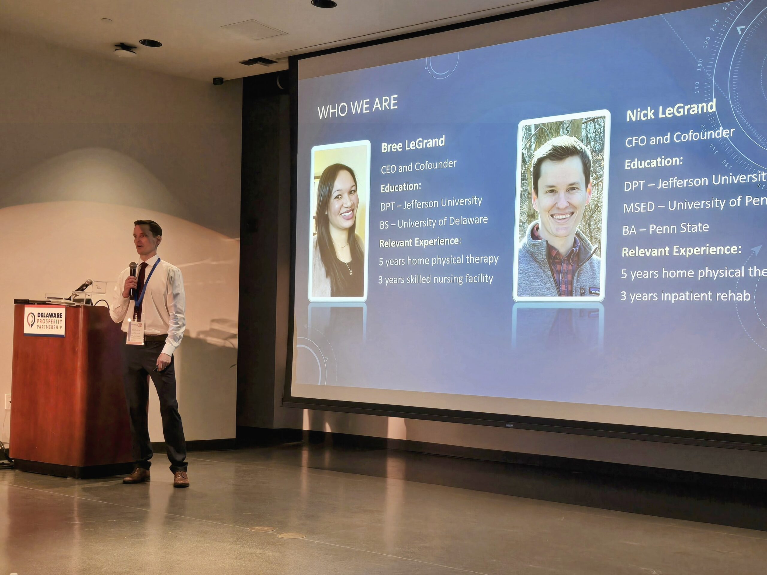 A man stands at a podium presenting a slide titled "Who We Are," which includes photos and bios of Bree LeGrand and Nick LeGrand, detailing their educational background and relevant experience.