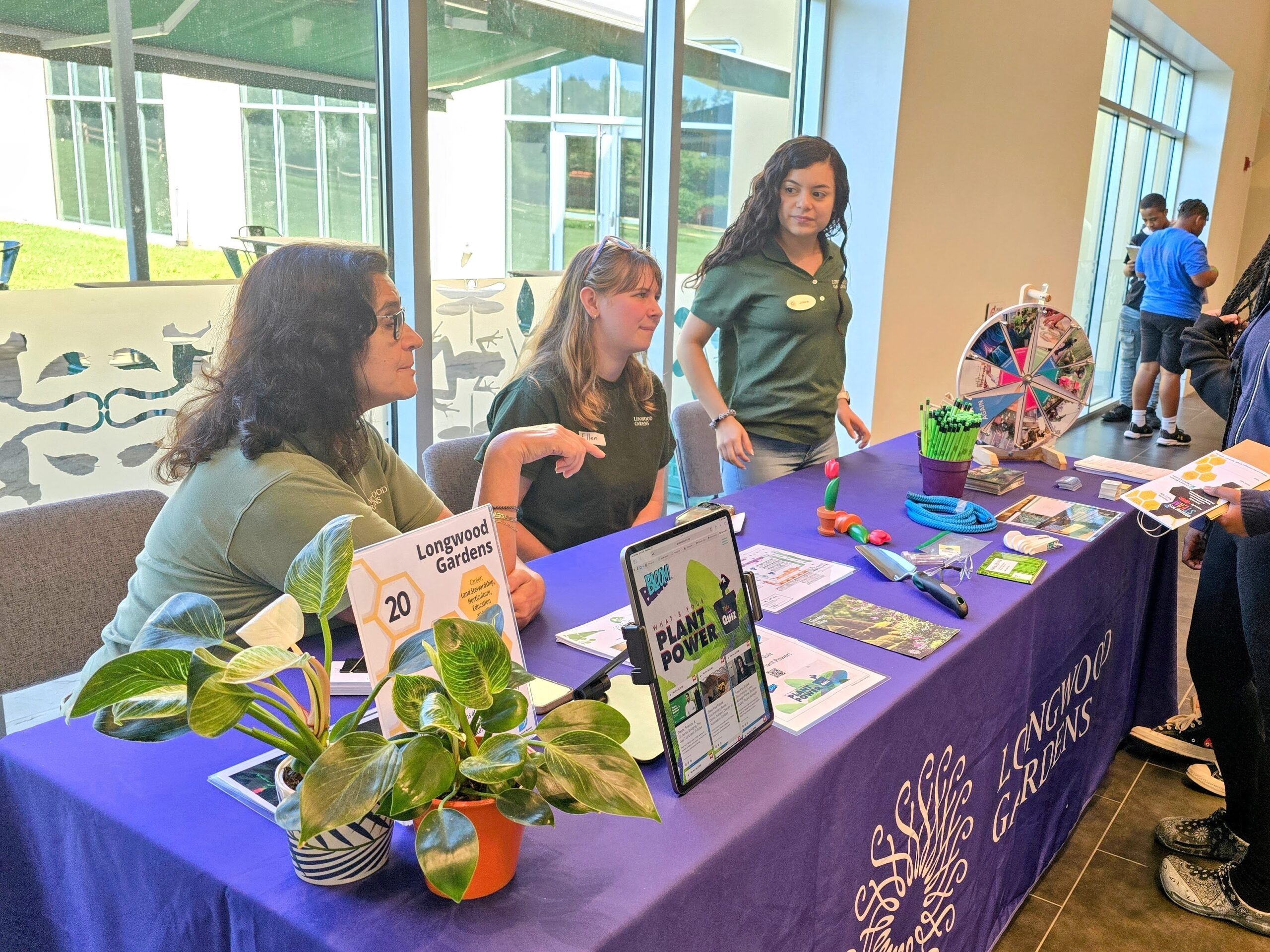 Three people at a Longwood Gardens booth talk with attendees. The booth features plants, brochures, a spinning wheel game, and promotional materials on a purple tablecloth.