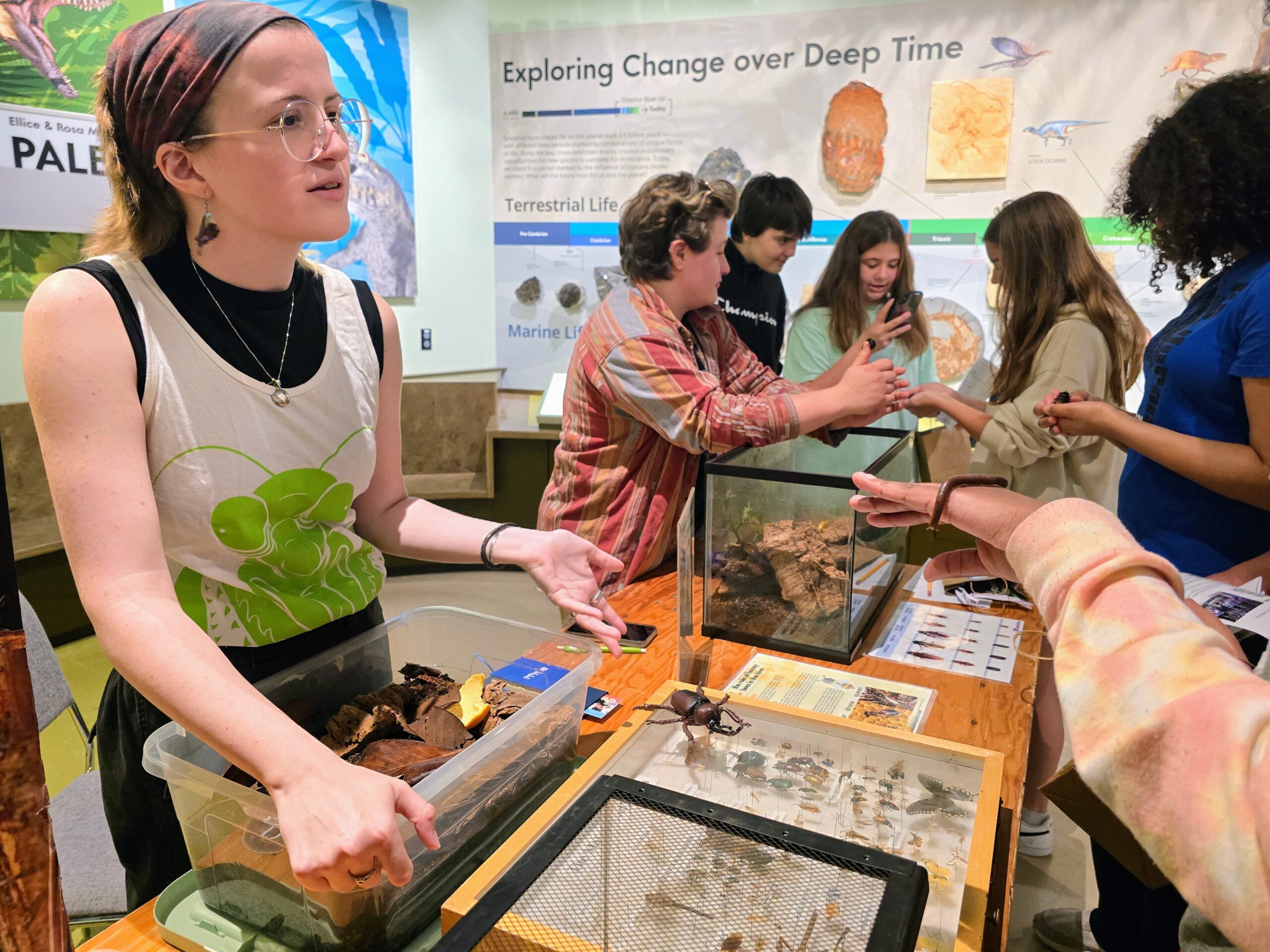 A person displays various specimens at a science exhibit while others observe and interact with the exhibits related to paleontology and environmental change.