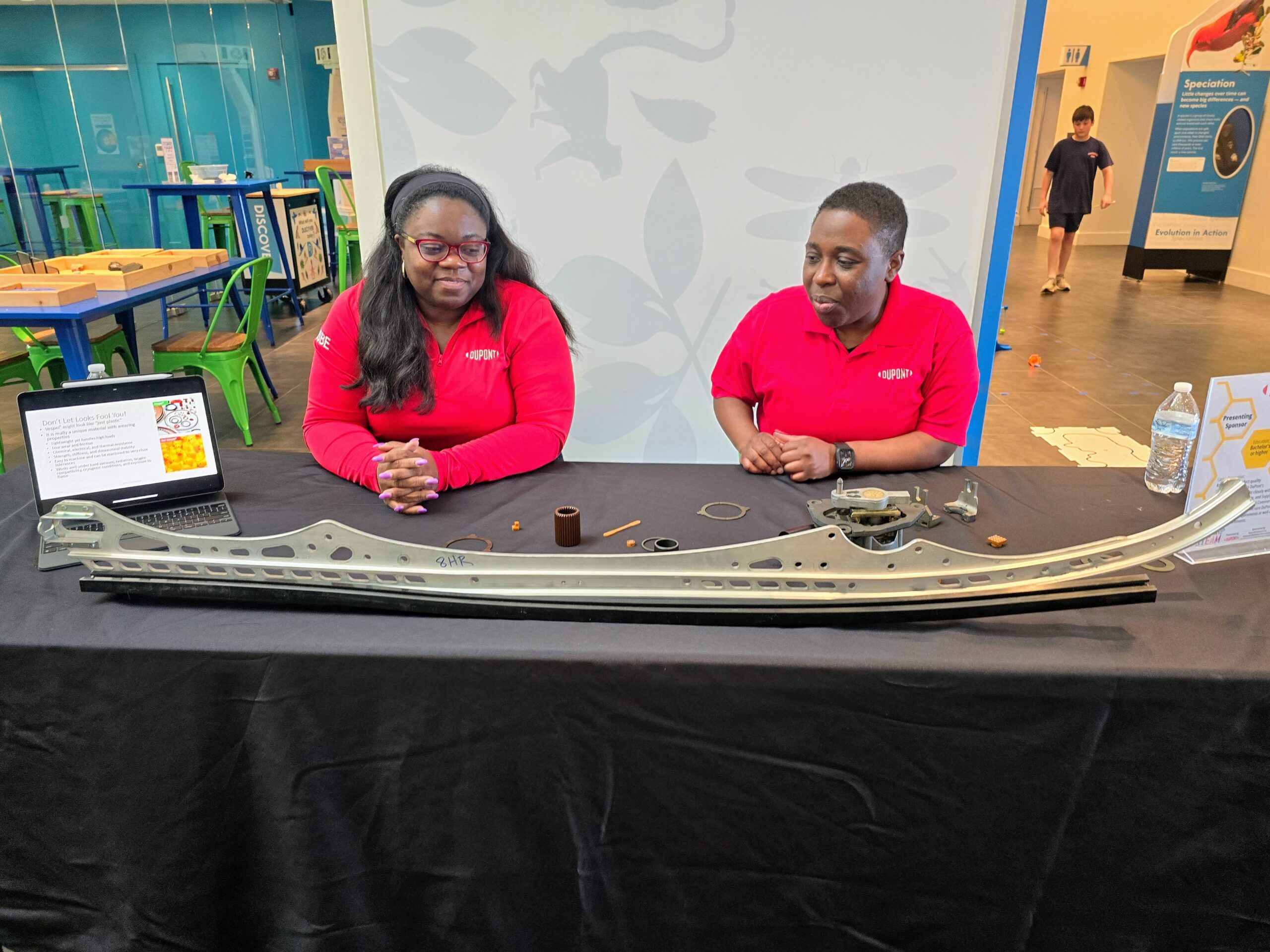 Two people in matching red shirts sit behind a table displaying aviation parts and information. A laptop and bottle are also on the table. The background features a science exhibit area with a few visitors.