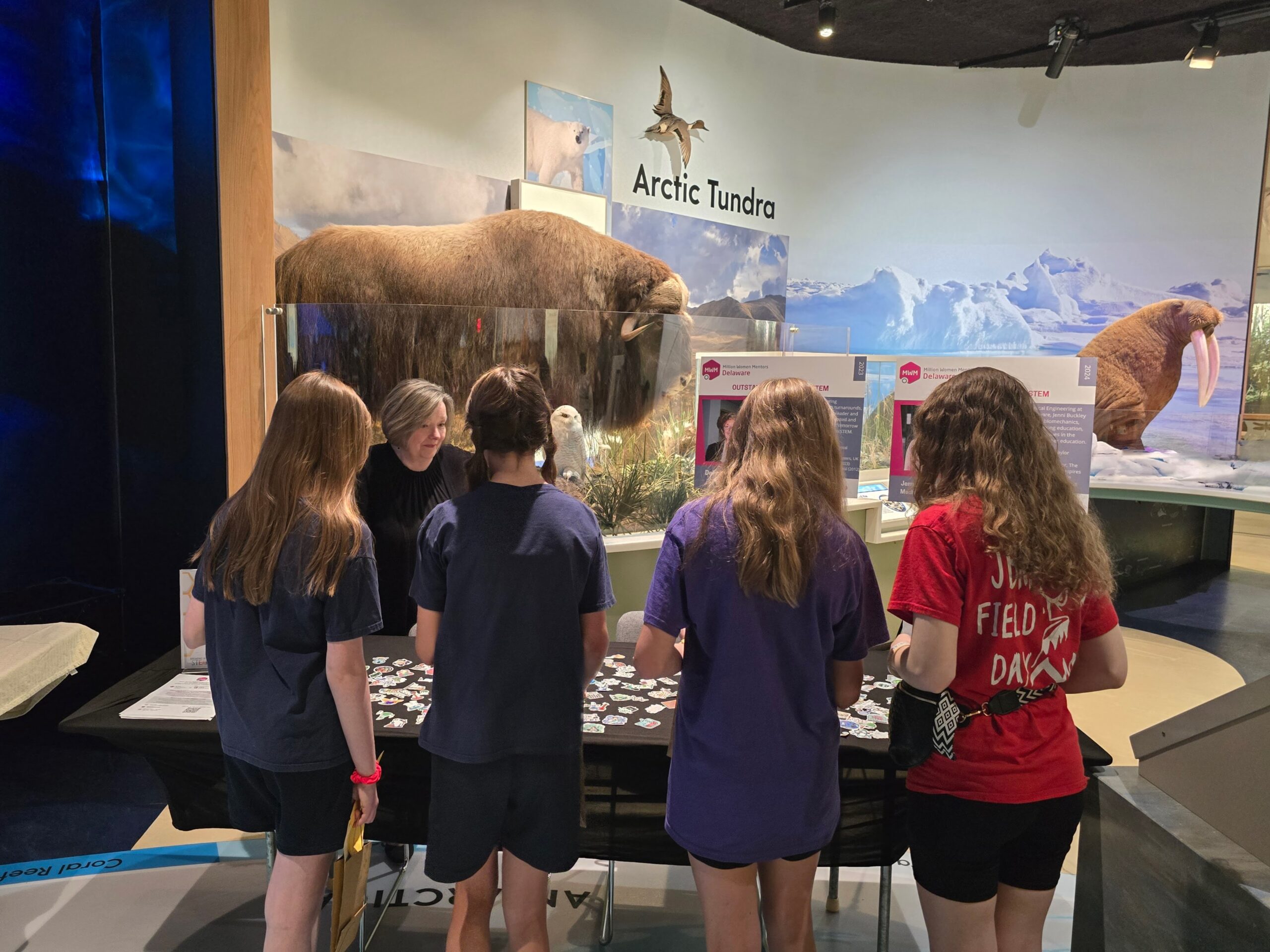 Group of children gathered around a table with badges, in an exhibit titled "Arctic Tundra" featuring a walrus display in the background.