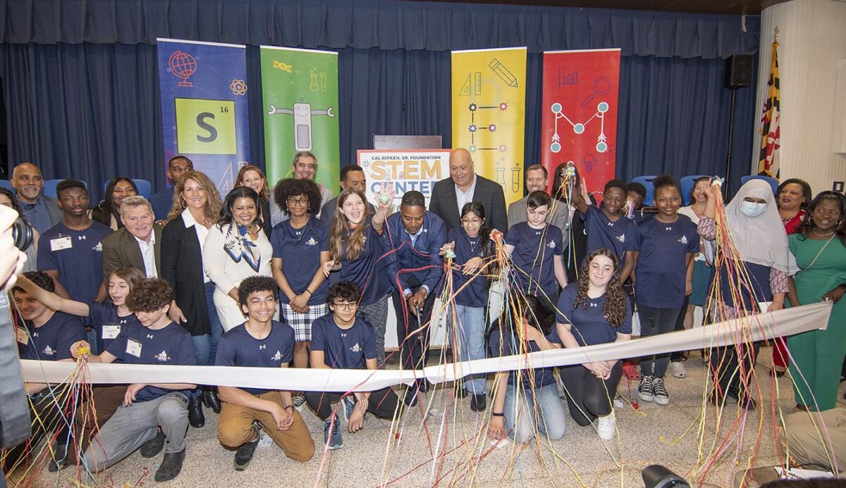 Children and adults in navy clothes pose by white ribbon and multicolored signs.