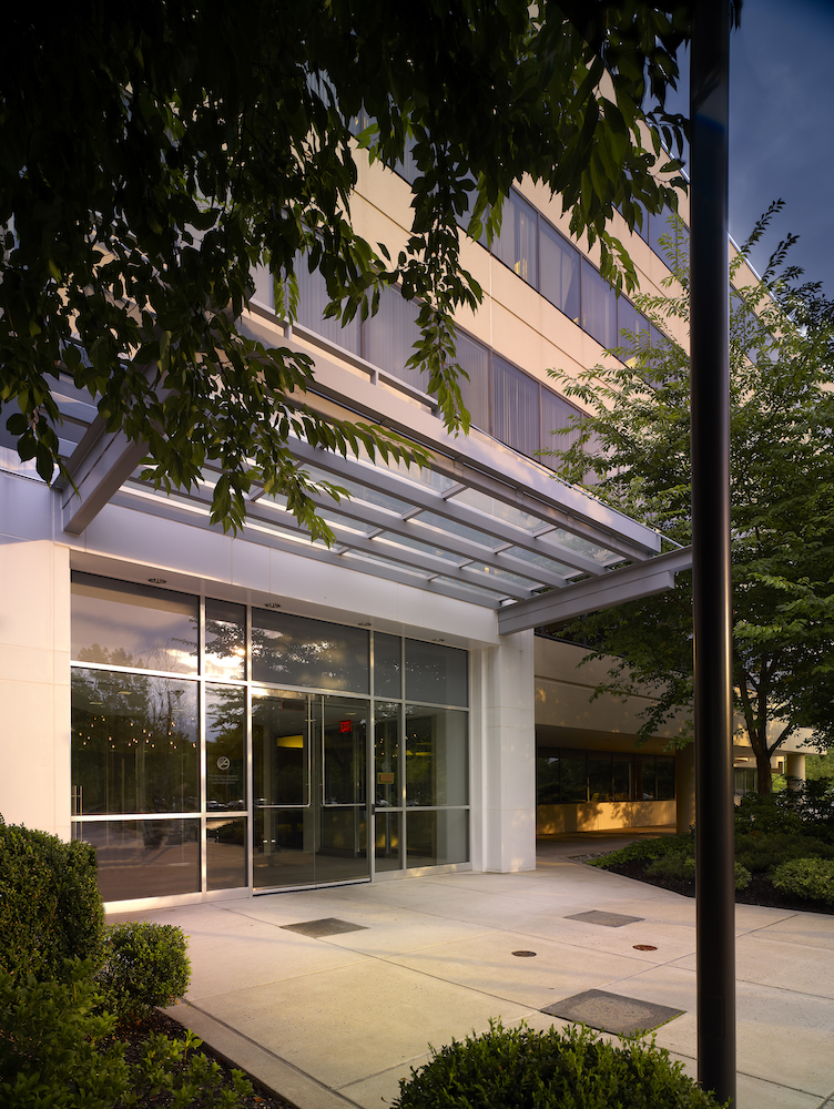 The entrance to an office building with trees and plants lining the sidewalk in front of large glass doors.