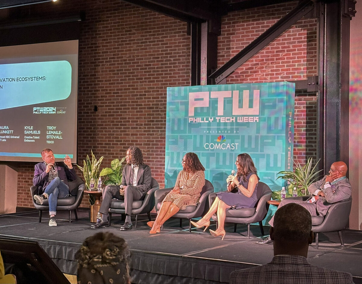 Panel of five individuals seated on a stage at Philly Tech Week, engaged in discussion with a large screen displaying event details in the background.