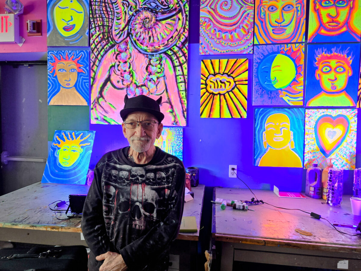 An older man wearing a black hat and a skull-themed sweater stands in front of a wall displaying brightly colored, psychedelic artworks.