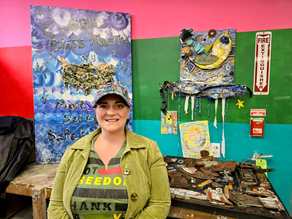A person wearing a hat and green jacket stands in front of colorful mixed-media art pieces on a wall. 