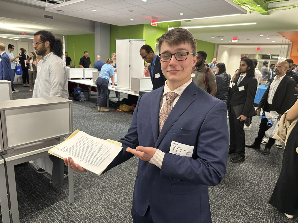 Young white man in a blue suit presenting a document at a crowded professional event.