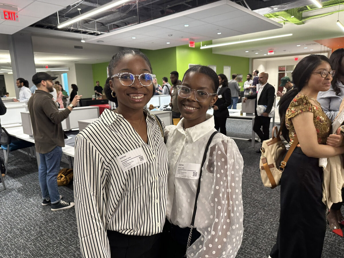 Two Black women smiling at a networking event, one in a striped shirt and the other in a polka dot blouse, both wearing glasses and name tags.