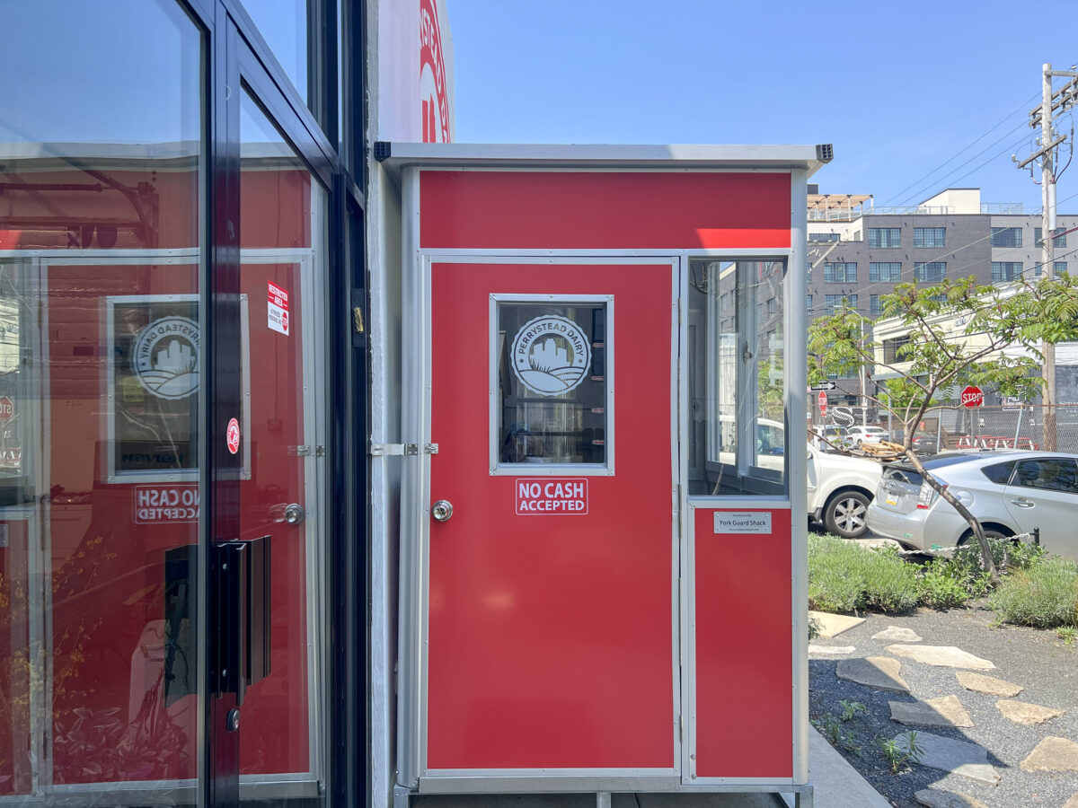 A small red booth with a "no cash accepted" sign, positioned near a street with visible cars and trees under a clear sky.