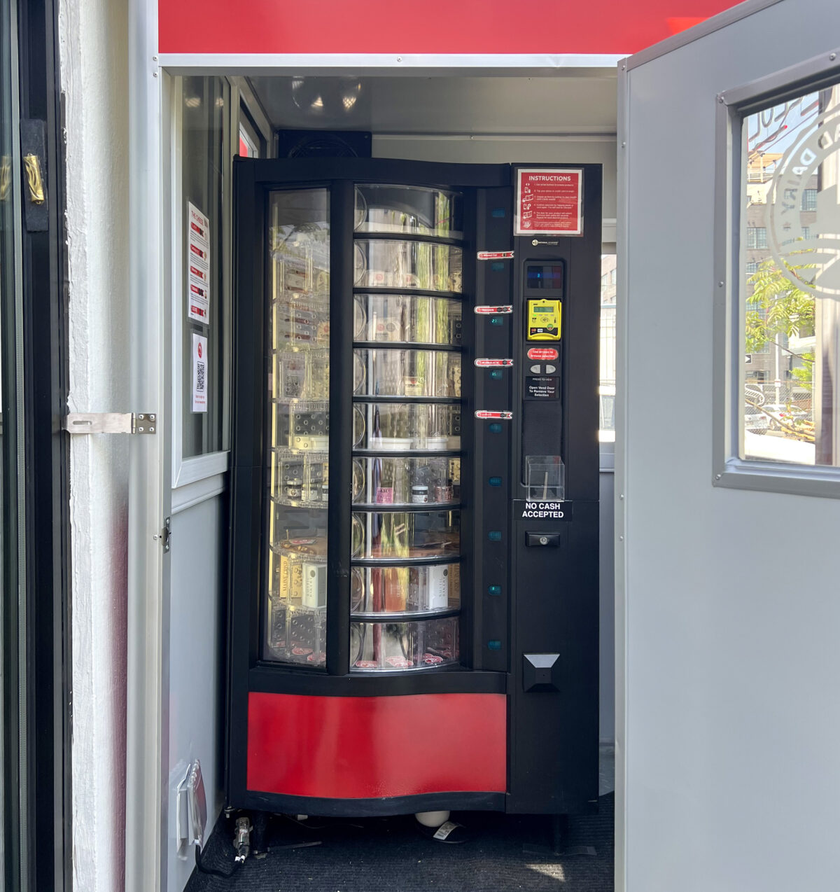 A red and black automated kiosk located in an outdoor setting, featuring a clear display of various items.