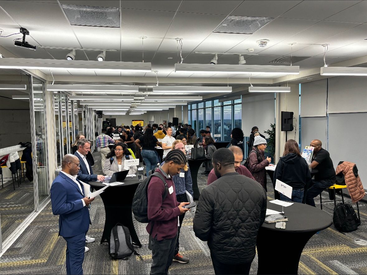 A large group of people gather in a large office room, talking to each other and reading books on papers, laptops, and phones.