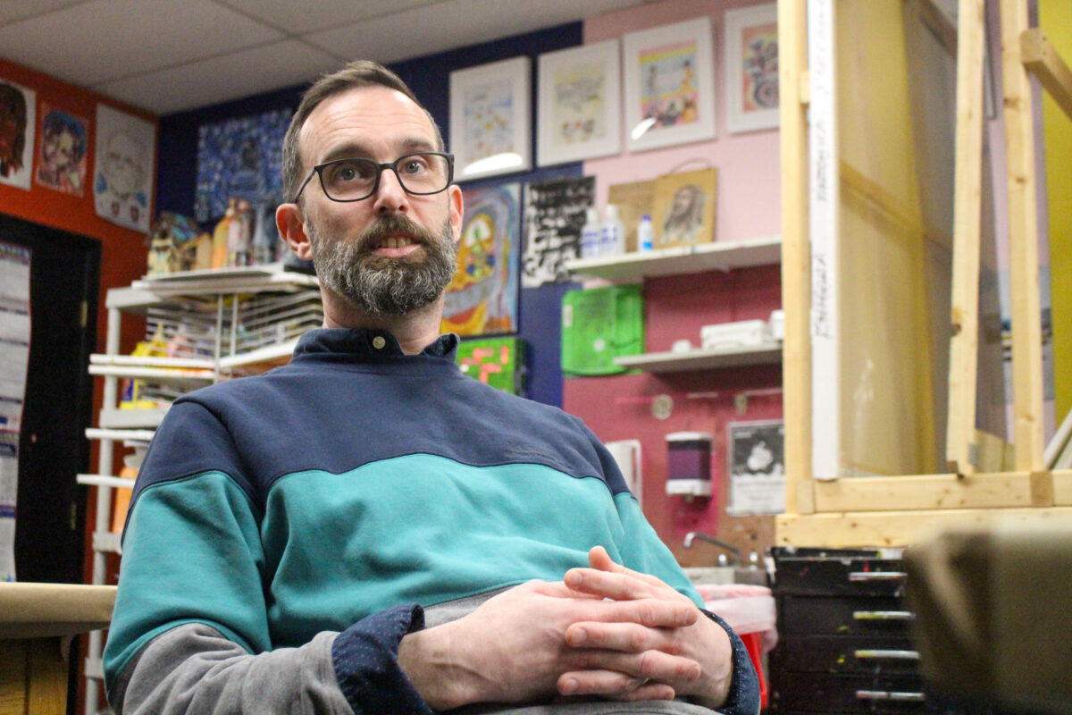 A man with glasses and a beard sitting in a colorful art room, looking slightly upwards with his hands clasped.