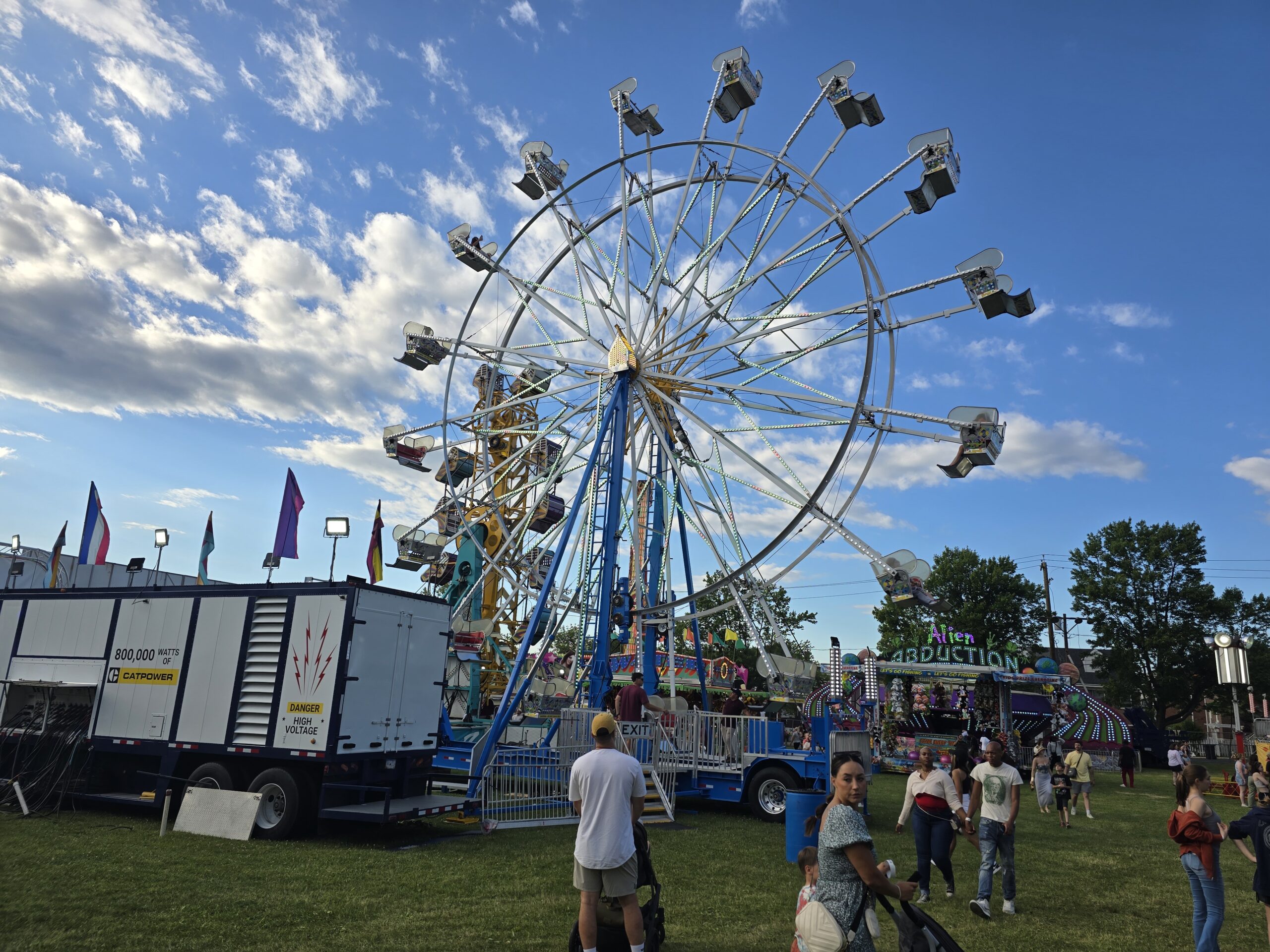 A large, white Ferris wheel with people in the seats is set up at a fairground. Other rides and attractions are visible in the background, with fairgoers walking and standing in the foreground.