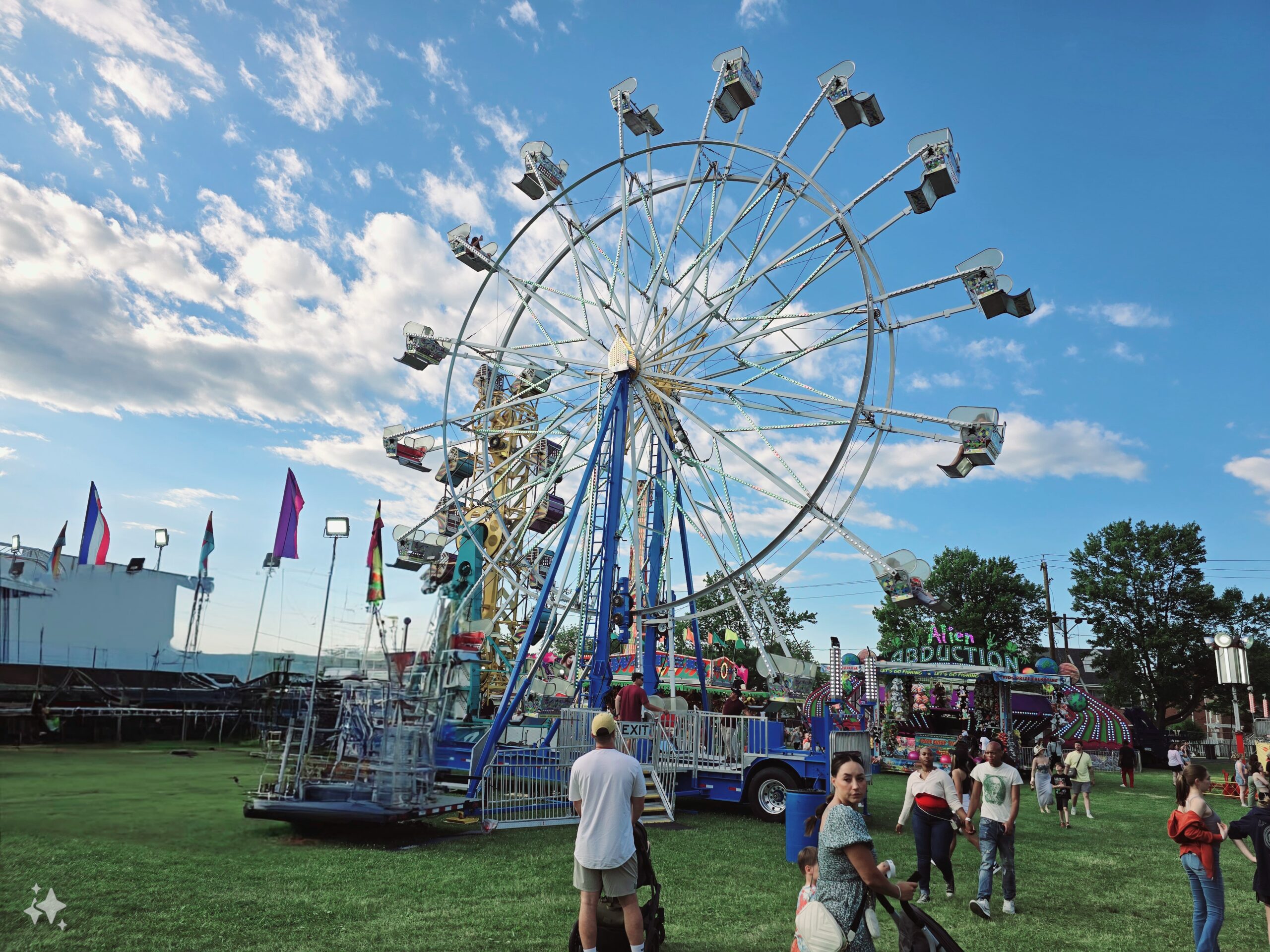 People gather near a Ferris wheel at an outdoor fair with colorful flags and a partly cloudy sky in the background.