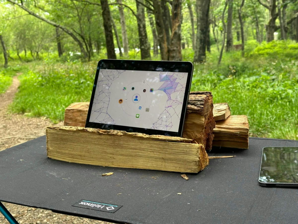 A tablet displaying a screen with icons rests on stacked firewood on a dark-colored table in a forest setting.