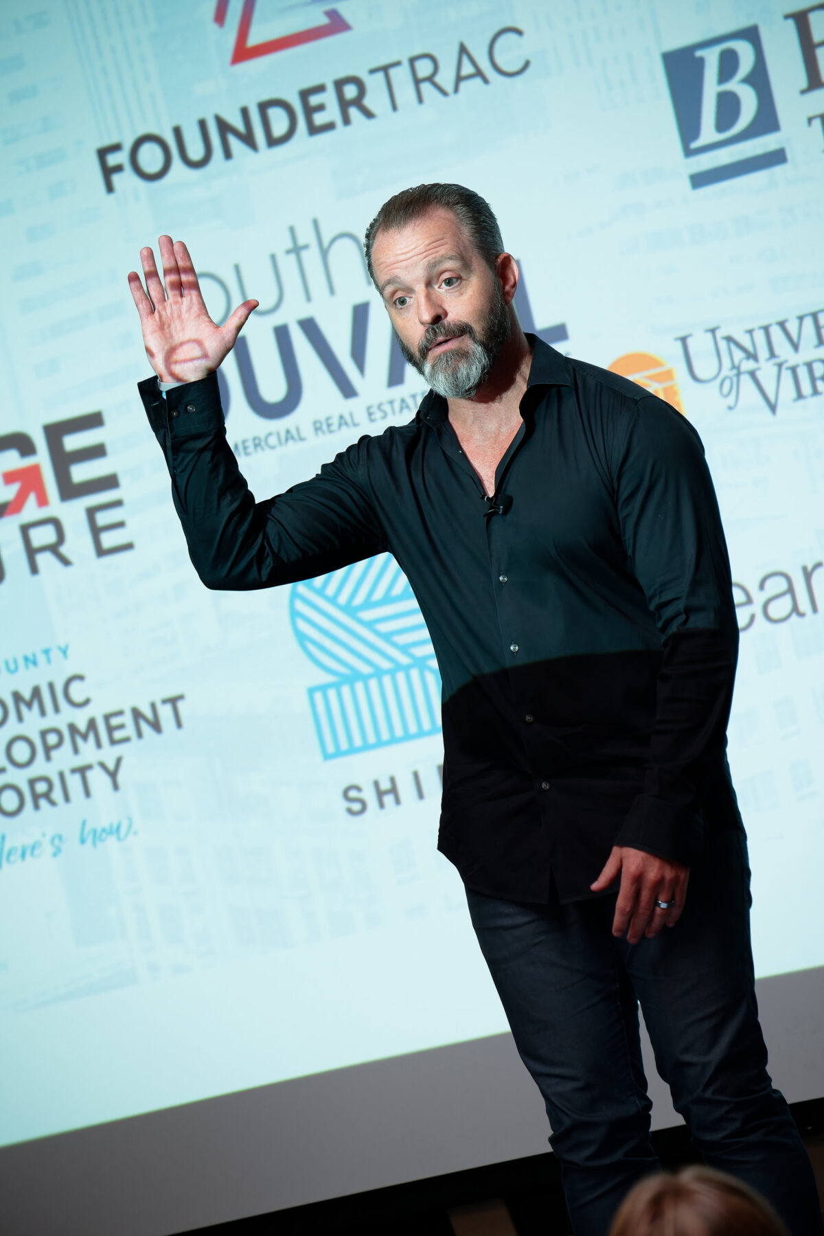 White bearded man in black shirt raises hand in front of projection screen