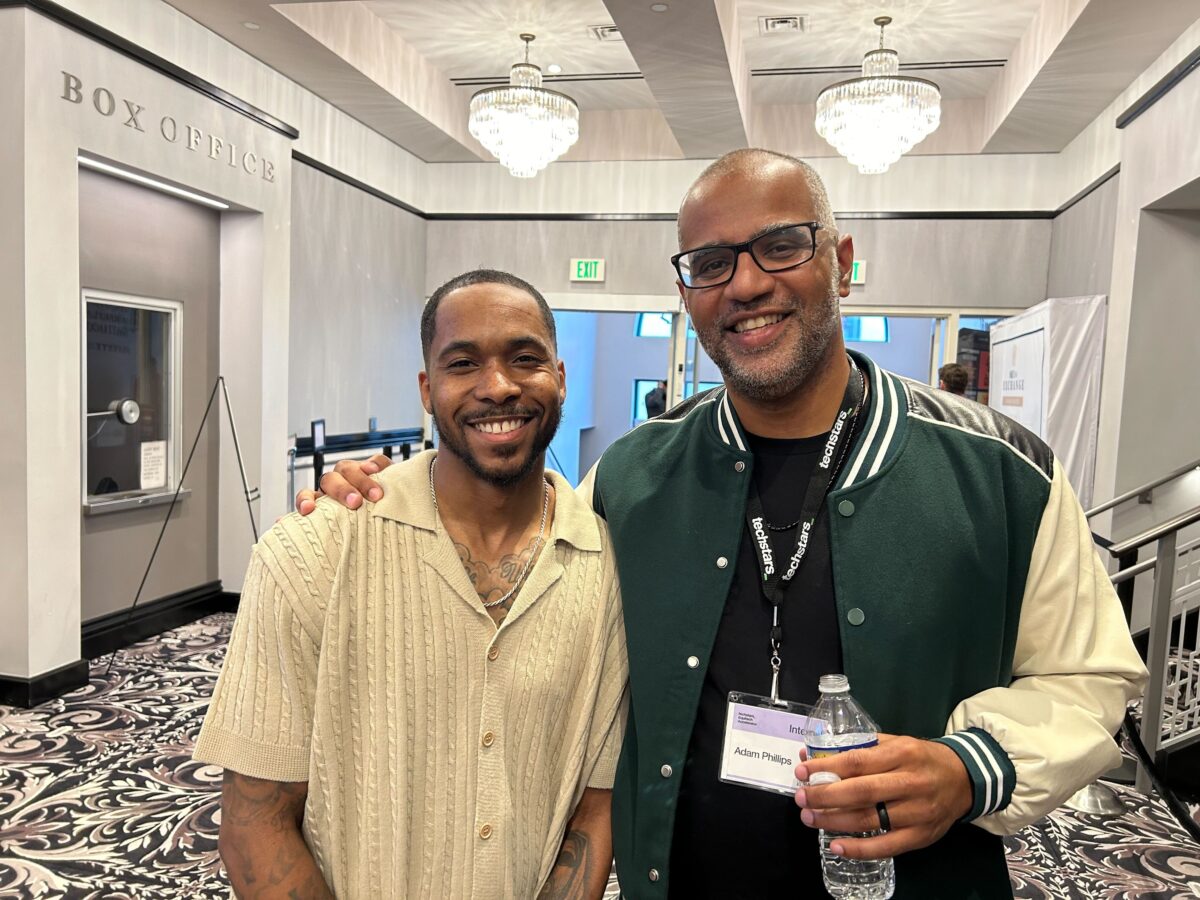 Two back men in beige and green clothing pose next to one another in room with black and brown decor.