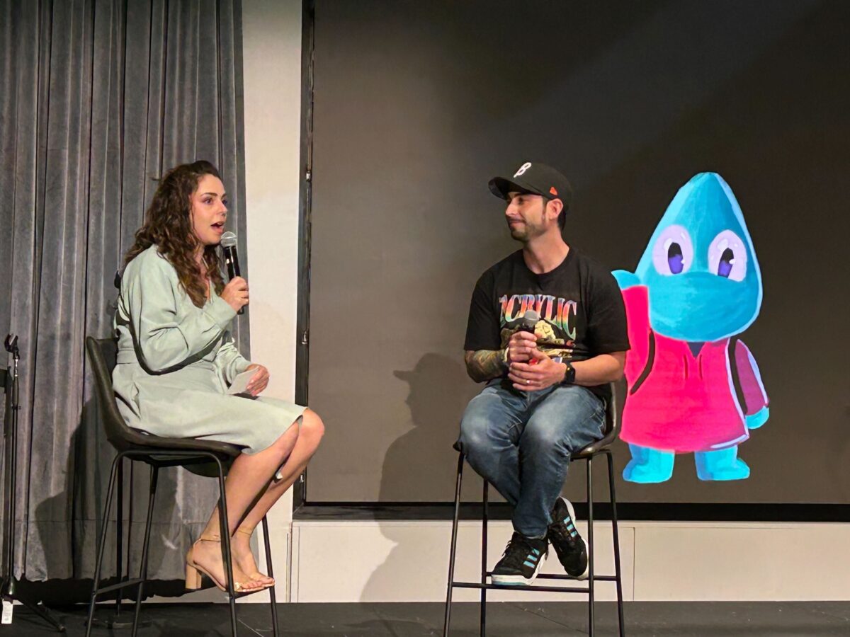 Woman in green dress speaks to man in black shirt and hat and blue jeans in front of screen with blue and pink icon.