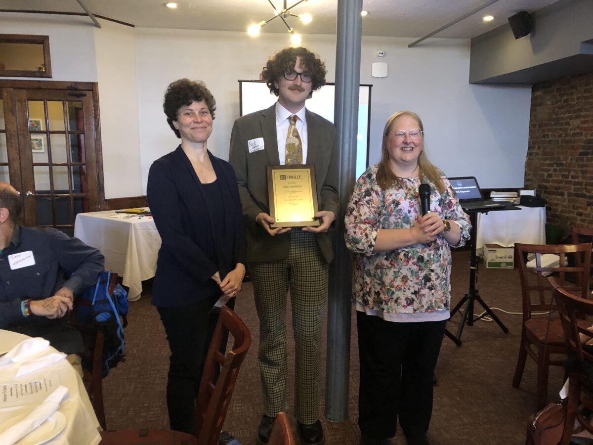 Three people standing together indoors; the person in the center holds a plaque. The person on the right is speaking into a microphone.